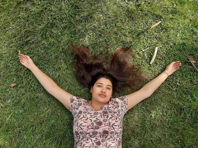 ZafiRouge's arms and legs are spread out on green grass, her brown hair fanned out, wearing a lace-patterned top, with a relaxed, content expression on her face.