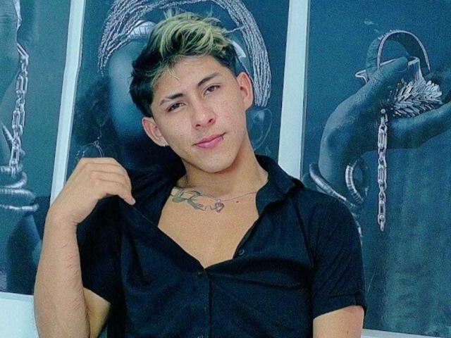 SoffiaVegga with a slight smirk, black shirt open, showing a tattoo on her chest, blond and black hair, holding shirt collar, in front of black-and-white wall art.