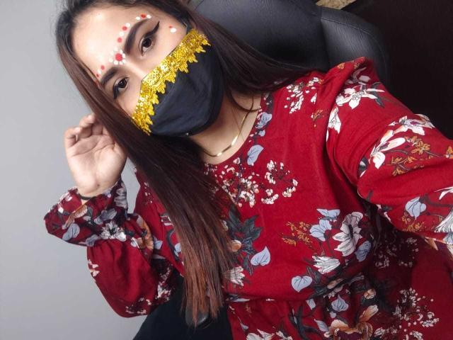 Dalia Dubey, con capelli castani, vestito floreale rosso, maschera nera, bordi in pizzo giallo, macchie bianche sul viso, seduta, con una mano vicino alla testa, che guarda la fotocamera, mostrando leggermente il seno..