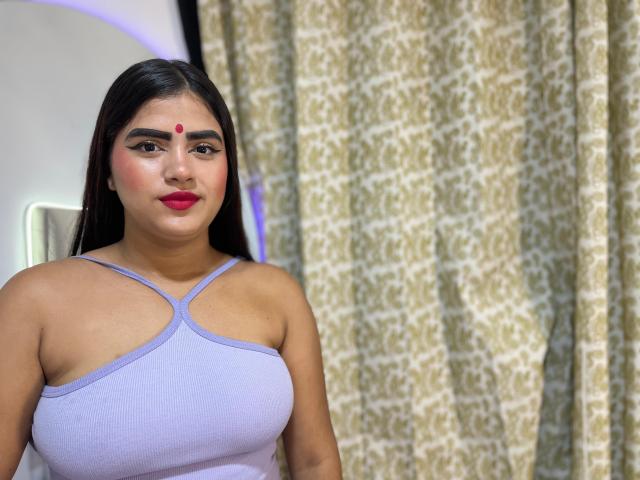 NayaYalitza with dark straight hair, wearing a lilac tank top showing her tits, red dot on her forehead, red lipstick, standing in front of patterned curtains.