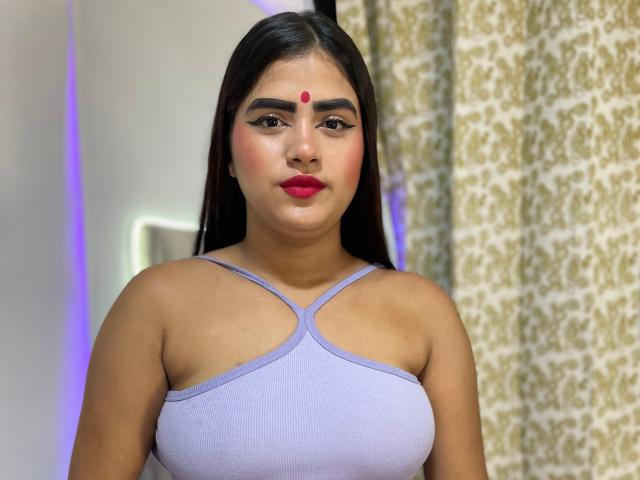 NayaYalitza with dark hair, red dot on forehead, purple tank top, showing her tits, red lipstick, and brown eyes, standing in front of patterned curtains.
