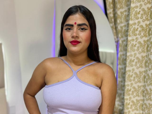 NayaYalitza with long dark hair, red dot on forehead, purple tank top, showing her tits, red lipstick, and a confident expression. She's standing indoors with a patterned curtain.