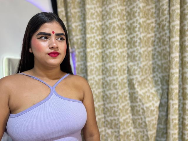 NayaYalitza with dark hair, red dot on forehead, wearing a lavender tank top, showing her big tits, red lipstick, and patterned curtains in the background.