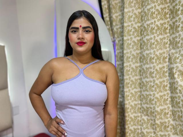 NayaYalitza with dark long hair, red dot on forehead, purple tank top, big boobs, red lipstick, standing with hands on hips, patterned curtain on right.