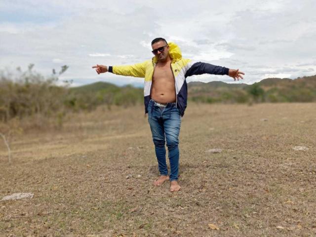 WolfineOne, shirtless with yellow and blue jacket, blue jeans, and sunglasses, stands barefoot with arms out, showing a flabby, tan belly in a grassy field.