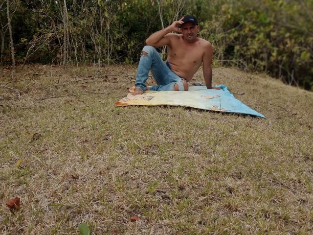 Shirtless WolfineOne with ripped jeans, black cap, and bare feet, sitting on a surfboard in grassy field, touching head with hand, looking relaxed.