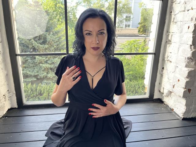 ElizabethSo with black wavy hair, dark lipstick, black dress, cleavage, and red nails, hand on belly, sitting on black wooden floor, white brick wall, window behind.