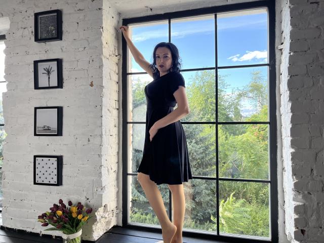 ElizabethSo stands in a black dress, her arm raised, showing her ass, against a white brick wall with a large window, green trees outside, and a flower vase below.