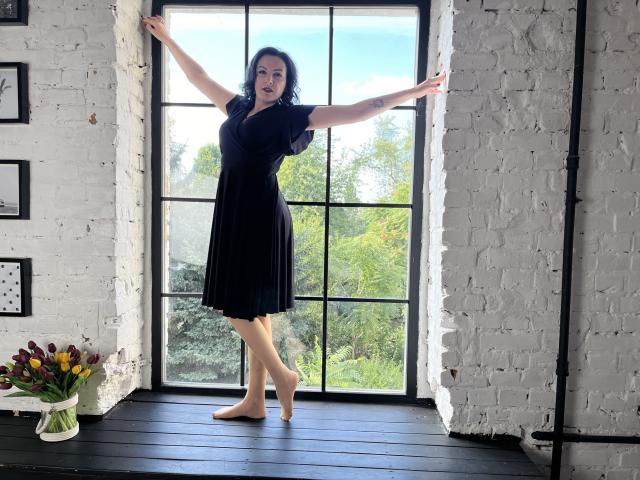 ElizabethSo stands barefoot on a black wooden floor, arms out, wearing a black dress, with white brick walls and a large window behind her. Her tits are visible.