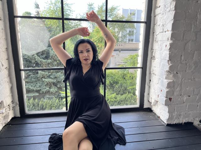 ElizabethSo sits on a black wooden floor, arms raised, wearing a black dress, exposing her tits, with white brick walls and a large window behind her.