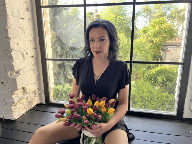 ElizabethSo with dark hair, black dress, holding bouquet of pink and yellow tulips, sitting on black window sill, white brick wall, green tree outside, looking slightly seductive.