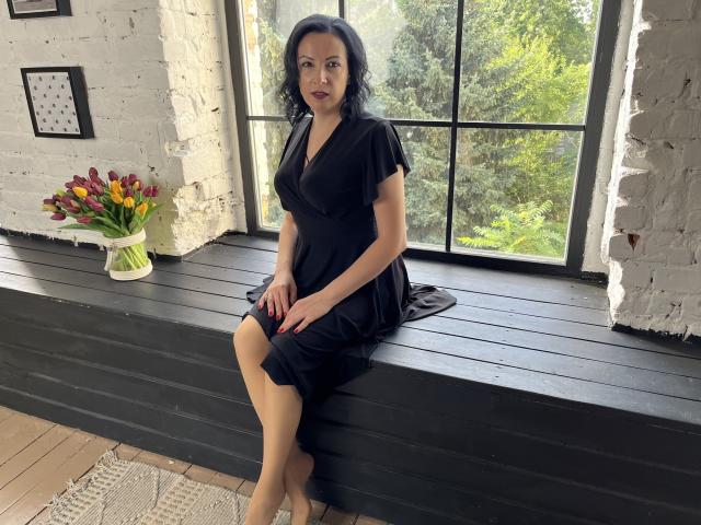 ElizabethSo with black dress, sitting on black window ledge, legs crossed, hands on lap, dark wavy hair, red lipstick, black brick wall, vase of colorful flowers on left.