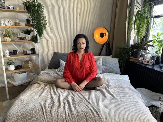 ElizabethSo with curly black hair, wearing a red button-up shirt, sits cross-legged on a bed in a cozy room, her hands resting on her knees, looking at the camera.