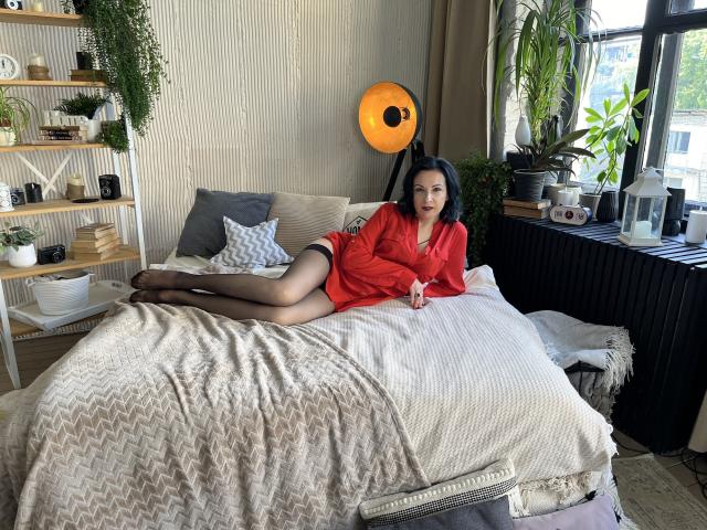 ElizabethSo, lying on a bed in a red shirt, black stockings, and no pants, showing her ass and pussy, with black hair and red lipstick, in a cozy room.
