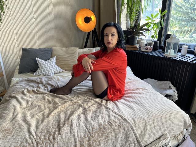 ElizabethSo with black hair, red shirt, and black stockings sits on a bed, legs crossed, showing her ass and black panties, with a lamp and plants in the background.