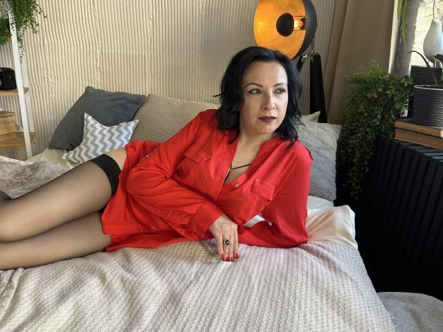 ElizabethSo with black hair, red shirt, black stockings, and visible ass, lying on a bed with a sexy look, dark lipstick, and red nails.