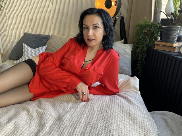 ElizabethSo with black hair, red shirt, and black stockings, lying on a bed, showing her tits, with a ring on her finger, and red nails, looking sexy.