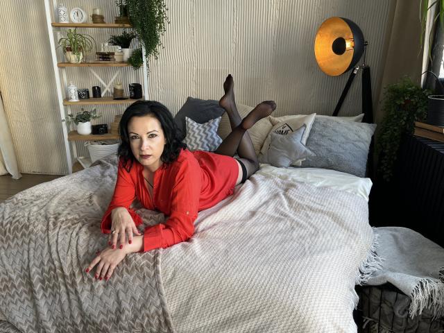 ElizabethSo with black hair, red shirt, black pantyhose, and red nails, lying on a bed, showing her ass, looking at camera, legs up, in a cozy, modern room.