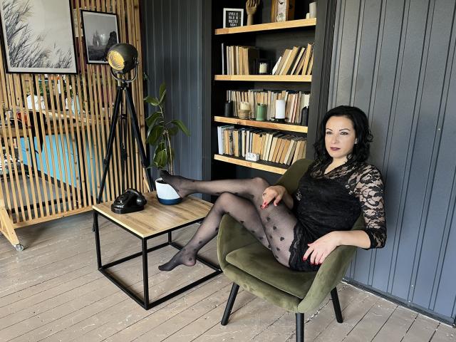 ElizabethSo with black lace dress, sheer black pantyhose, and polka dots, sitting in green chair, legs crossed, showing her pussy, black boots on wooden table, gray wall.