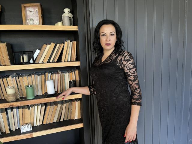 ElizabethSo with black lace dress, medium tits, black wavy hair, dark lipstick, leaning on bookshelf, grey wall, wooden shelves with candles and books, standing slightly to the right.