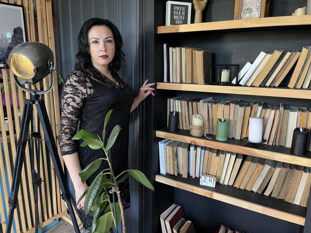 ElizabethSo with black lace dress, standing next to wooden bookshelves, green plant, and a tripod lamp, has her hand on the shelf, showing off her tits.