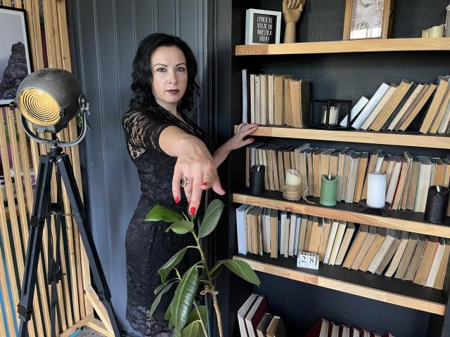 ElizabethSo with black lace dress, red nails, pointing at bookshelf, dark hair, standing next to green plant, studio light on tripod, bookshelves, black wall, wooden shelves, books, candles.