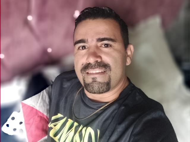 WolfineOne has a medium build, dark hair, goatee, and is smiling at the camera wearing a black shirt with a gold necklace. His eyes are brown.