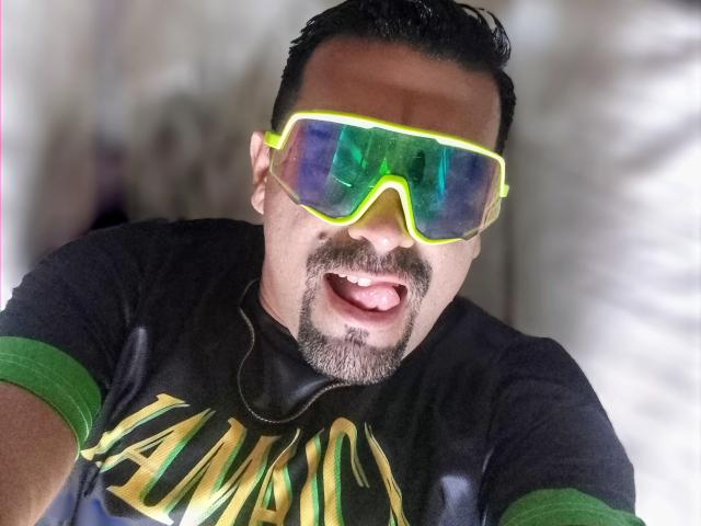 WolfineOne wearing neon green reflective sunglasses, black t-shirt with green sleeves, and a goatee, with tongue out, looking up at the camera.