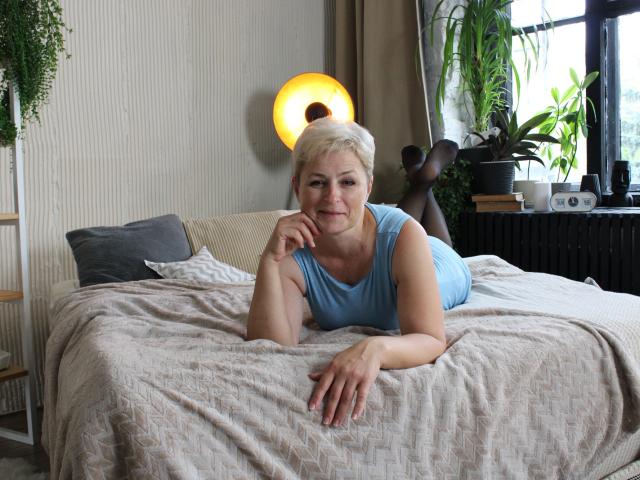 EmiliaGree with short blonde hair, lying on a bed in a blue dress, resting on her stomach, legs up, smiling, right hand on chin, light skin, black stockings.