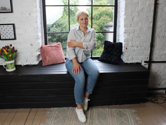 EmiliaGree with short blonde hair, crossed arms, wearing a grey shirt, blue jeans, and white sneakers, sitting on a black wooden bench with pink and black pillows.