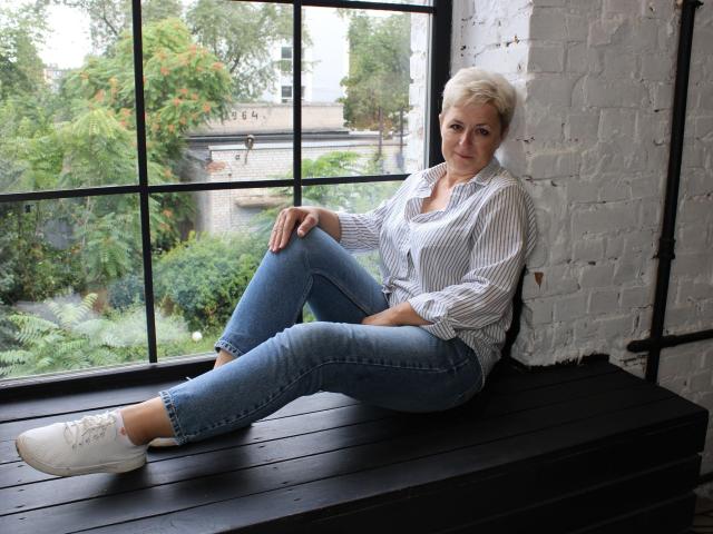 EmiliaGree with short gray hair, striped shirt, blue jeans, and white sneakers, sitting on black wooden bench, leaning against white brick wall, legs crossed, smiling.