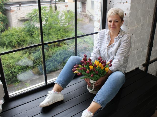 EmiliaGree sits on a black wooden window seat, wearing a striped shirt, blue jeans, and white sneakers, holding a bouquet of red, yellow, and purple tulips.