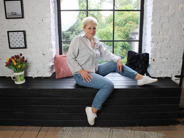 EmiliaGree with short blonde hair, wearing a grey striped shirt, blue jeans, and white sneakers, sitting on a black wooden bench with pink and black pillows, and white brick wall.