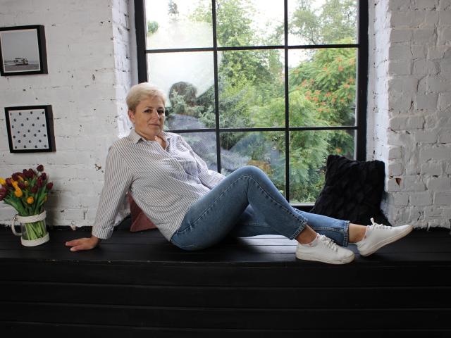 EmiliaGree with short gray hair, wearing a striped shirt and blue jeans, sits on a black wooden windowsill, legs outstretched, white sneakers on, black pillow beside her.