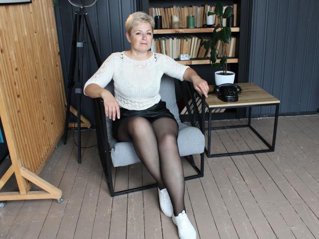 EmiliaGree, short blonde hair, white lace top, black mini skirt, black pantyhose, white sneakers, sitting on grey chair, wooden floor, bookshelf, potted plant, tripod in background.