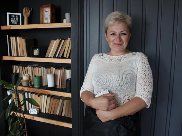 EmiliaGree stands with short blonde hair, wearing a white lace top, black pants, holding a book, smiling, next to a wooden bookshelf with books and decor.