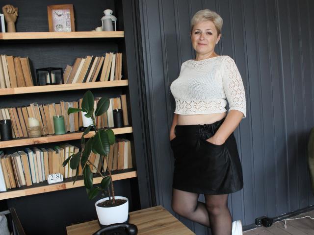 EmiliaGree with short blonde hair, white lace crop top, black skirt, hands in pockets, standing, black wall, wooden bookshelf, potted plant, gray floor, and wooden table.