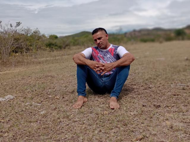 WolfineOne sits barefoot on dry grass, legs bent, hands resting on knees, wearing a white shirt with red and blue patterns, and blue jeans, looking downward.
