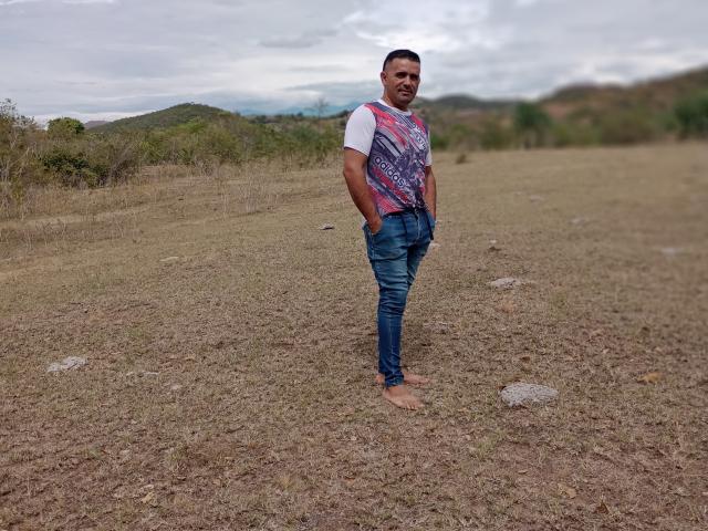 WolfineOne, barefoot, standing on dry grass, wearing blue jeans, white shirt with pink and purple pattern, hands in pockets, short dark hair, slightly smiling, cloudy sky, outdoors.