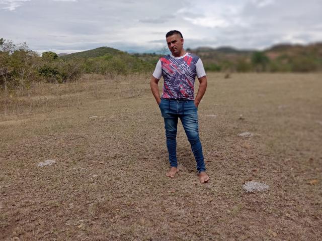 WolfineOne stands barefoot on dry grass, hands in jeans pockets, wearing a white shirt with red and blue graphic, and blue jeans, with a slight smile.