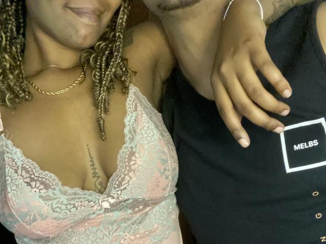 BrazilCouple with curly brown hair, wearing a pink lace bra, showing small tits, arm around a guy in a black shirt, hand on his chest, gold chain.