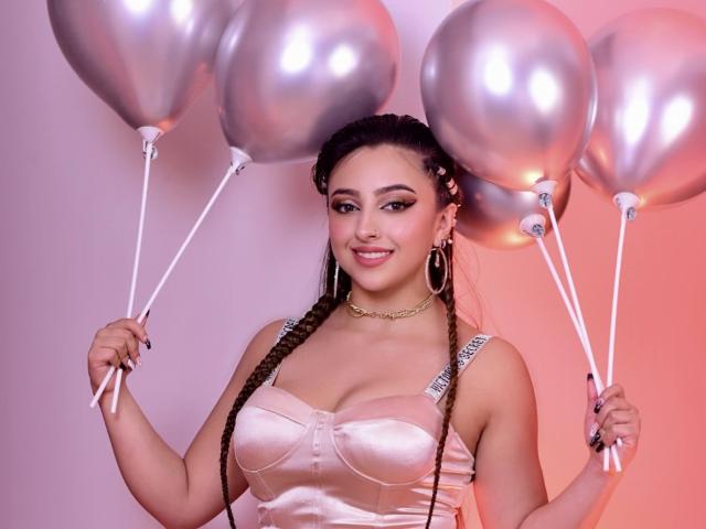 KylieCharm with shiny silver balloons, holding two in each hand. She's got long braids, big tits in a pink satin dress, and a cute smile. She's wearing hoop earrings.