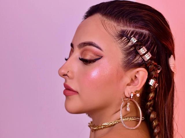 KylieCharm has glossy brown hair with diamond hairpins, wearing bold black eyeliner, pink blush, and gold hoop earrings. Her nose is pierced, and she's looking down.