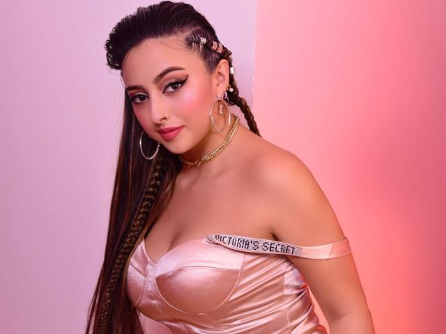 KylieCharm a de longs cheveux bruns tressés, elle porte une robe rose brillante avec une épaule dénudée, un collier en chaîne dorée et des boucles d'oreilles créoles, avec l'étiquette "Victoria's Secret" sur ses seins..