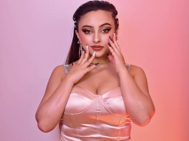 KylieCharm, con las manos en el rostro, lleva puesto un top rosa brillante de satén que deja ver sus grandes senos. Tiene uñas largas, un collar de oro y un fondo que va del rosa al naranja..