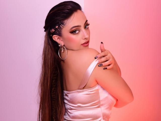 KylieCharm met lang bruin haar, een vlecht aan de zijkant, draagt een glanzend roze satijn top, grote oorringen en zwarte nagellak, kijkt over haar schouder, met haar hand op haar schouder..
