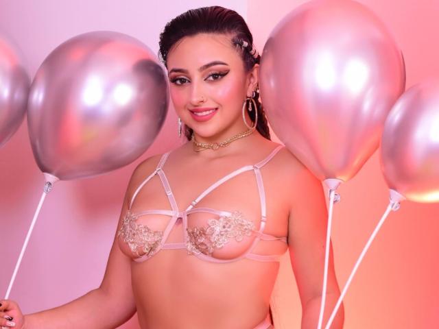 KylieCharm with shiny silver balloons, wearing a pink lace bra, has puffy tits, pink makeup, hoop earrings, and a gold choker, smiling at the camera.