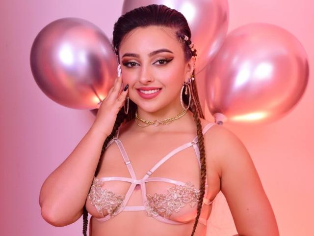 KylieCharm avec de longues tresses, portant de la lingerie en dentelle rose, de grandes boucles d'oreilles, une chaîne en or, des ballons roses, souriante, une main sur la joue, éclairage rose et violet. Seins, mignonne, sexy..