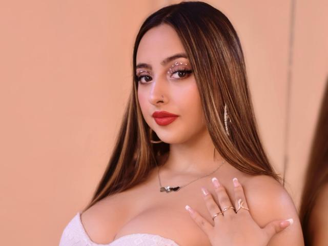 KylieCharm, con lunghi capelli castani, rossetto rosso e top bianco senza spalline, che mette in mostra il suo seno prosperoso e tiene un anello sulla sua mano curata, indossando una collana..