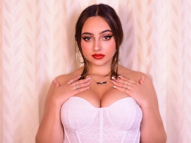 KylieCharm, avec ses cheveux bruns foncés, son rouge à lèvres rouge et son caraco en dentelle blanche, les mains sur ses seins, portant des bagues et un collier, regardant l'appareil photo avec une expression confiante..